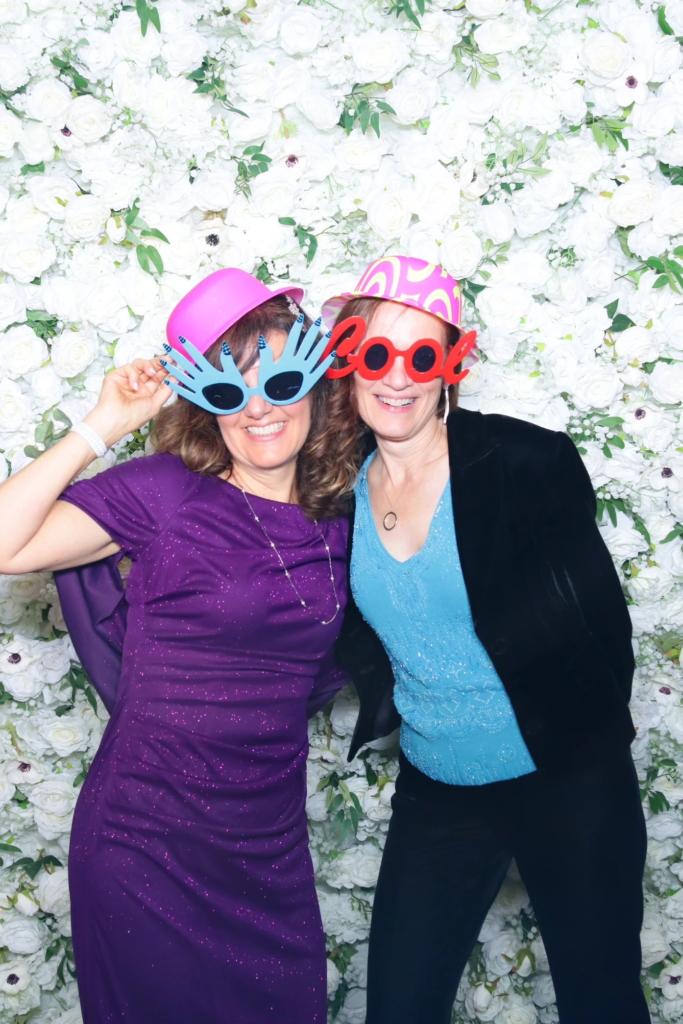Two women in colorful hats and novelty sunglasses posing in front of a white floral wall backdrop.