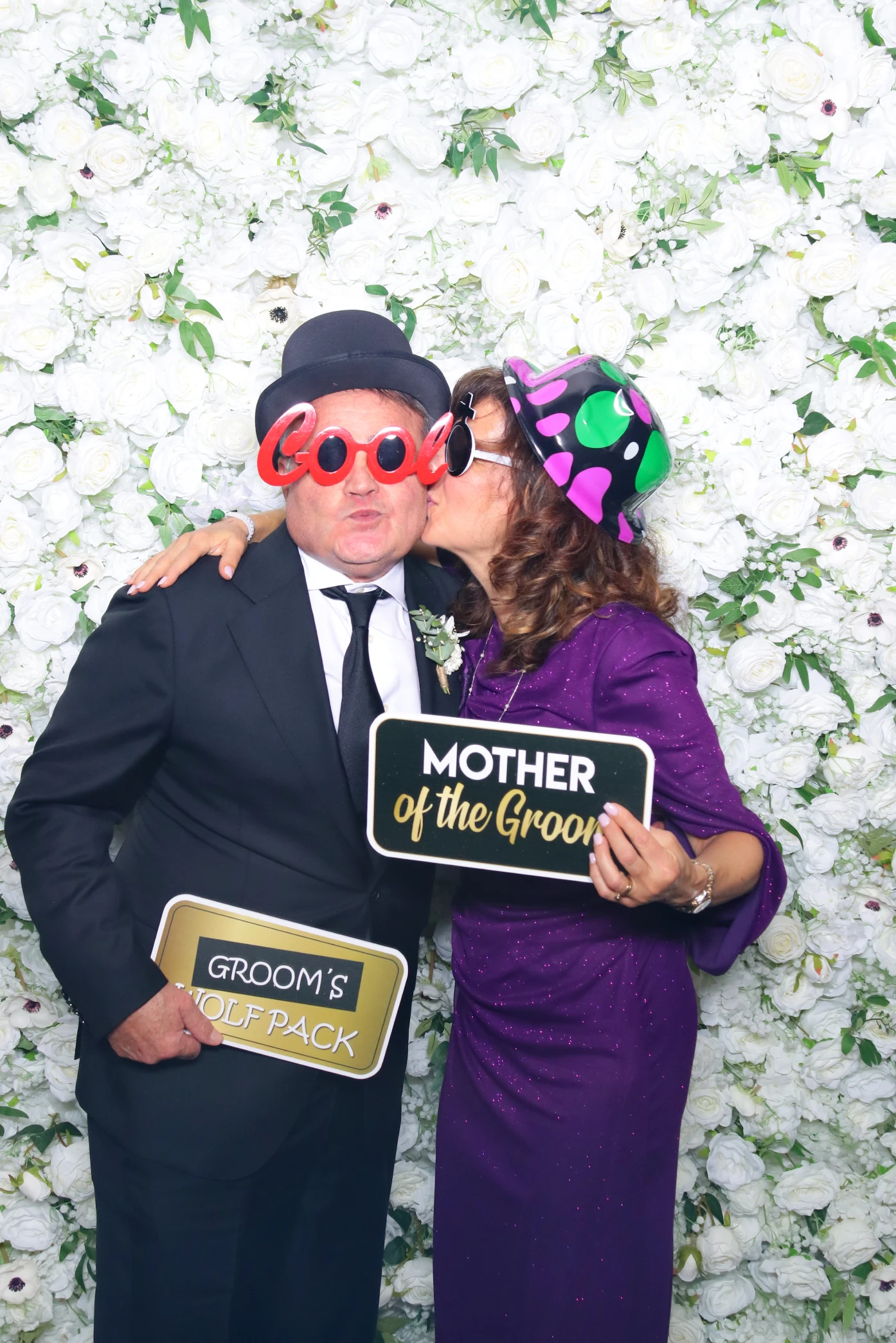 Man in a black suit and bowler hat wearing red 'cool' glasses and a woman in a purple dress and black hat with green and pink spots kissing his cheek, holding signs saying 'Groom's Wolf Pack' and 'Mother of the Groom' in front of a white rose flower wall.