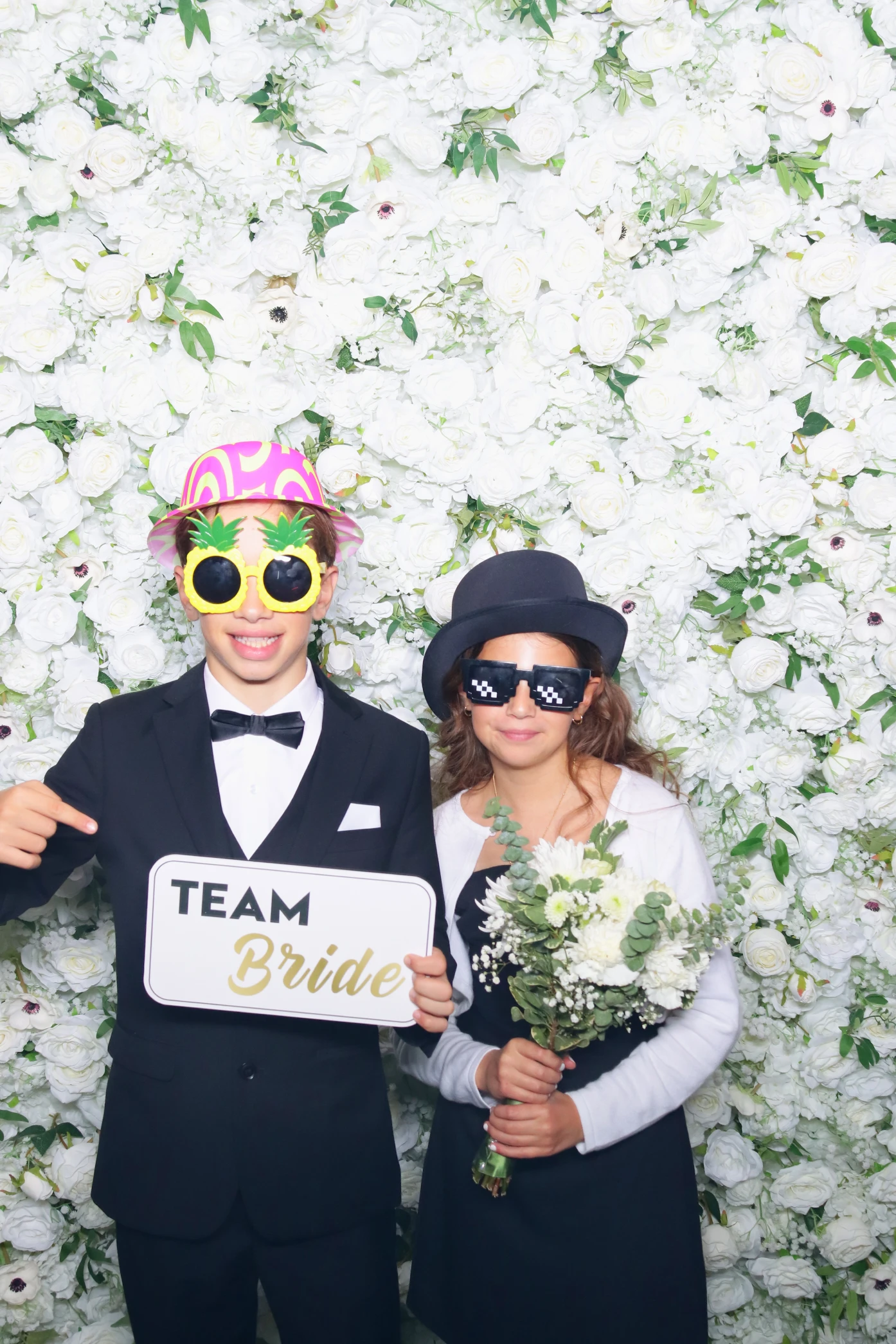 Two children in costume posing in front of a white floral wall, one holding a 'Team Bride' sign and the other holding a bouquet of white flowers.