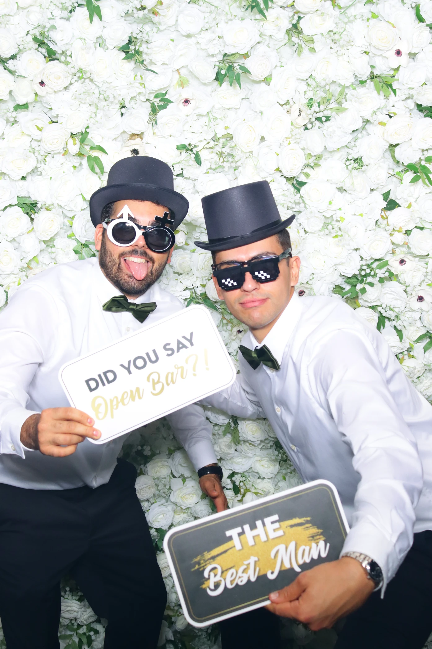 Two men in white shirts, black bow ties, and top hats posing in front of a white floral backdrop holding signs that say 'Did you say Open Bar?!' and 'The Best Man'.