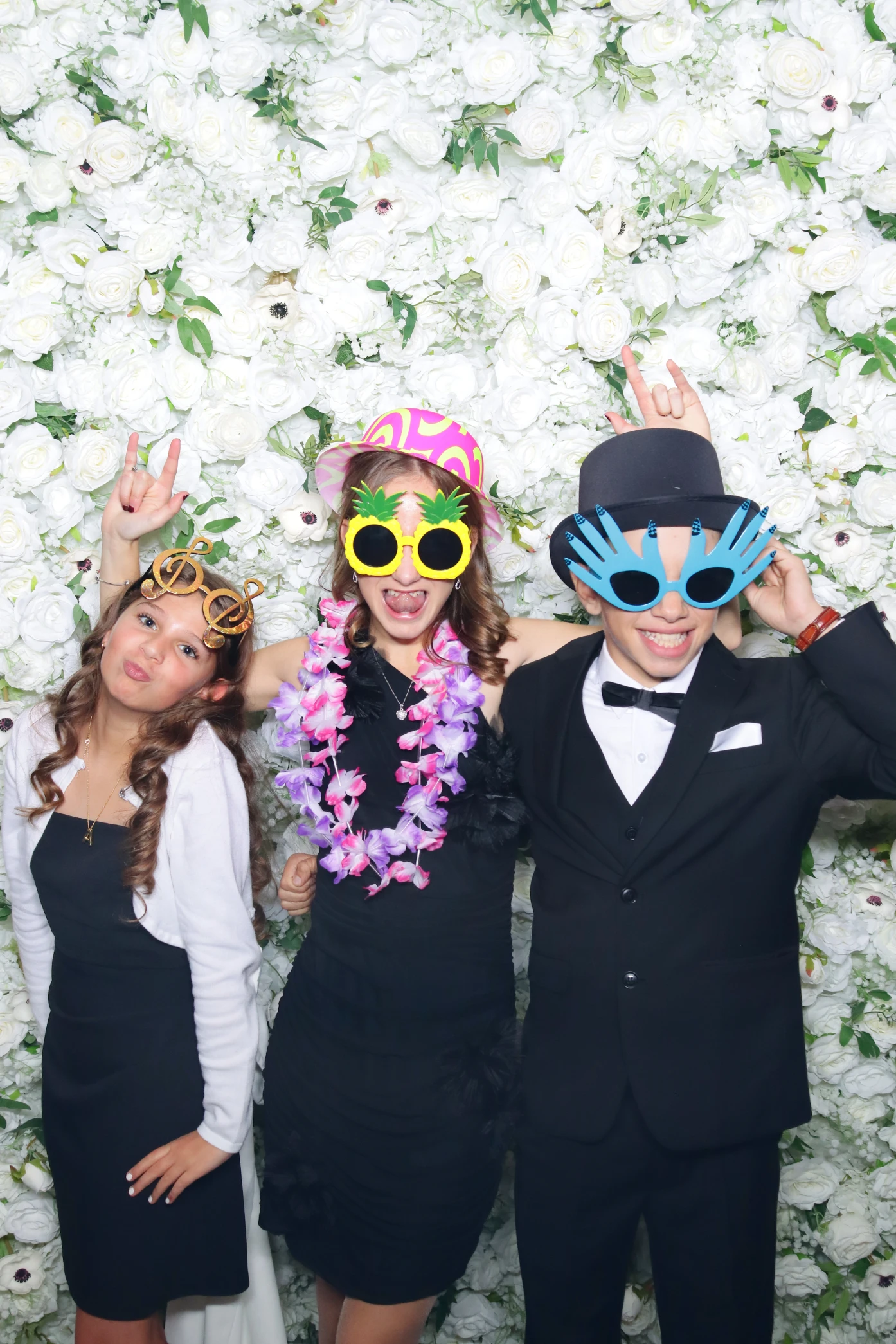Three young people in formal attire posing playfully in front of a white floral wall, wearing quirky oversized sunglasses and hats.