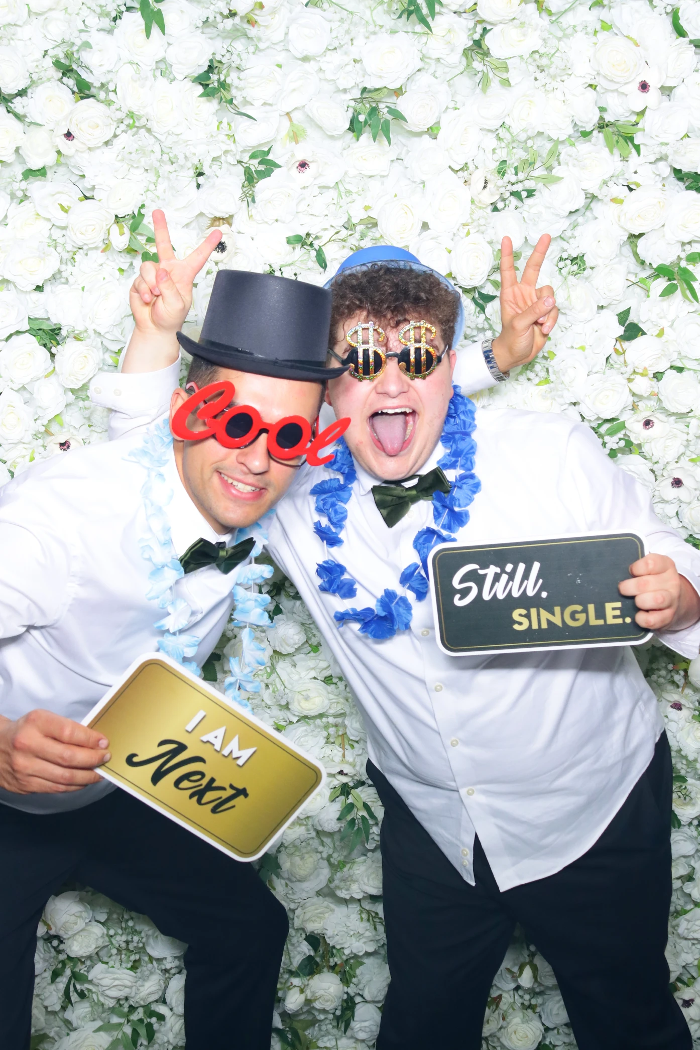 Two men in white shirts and bow ties posing in front of a white floral wall, wearing novelty glasses and holding signs that say 'I AM Next' and 'Still. SINGLE.'