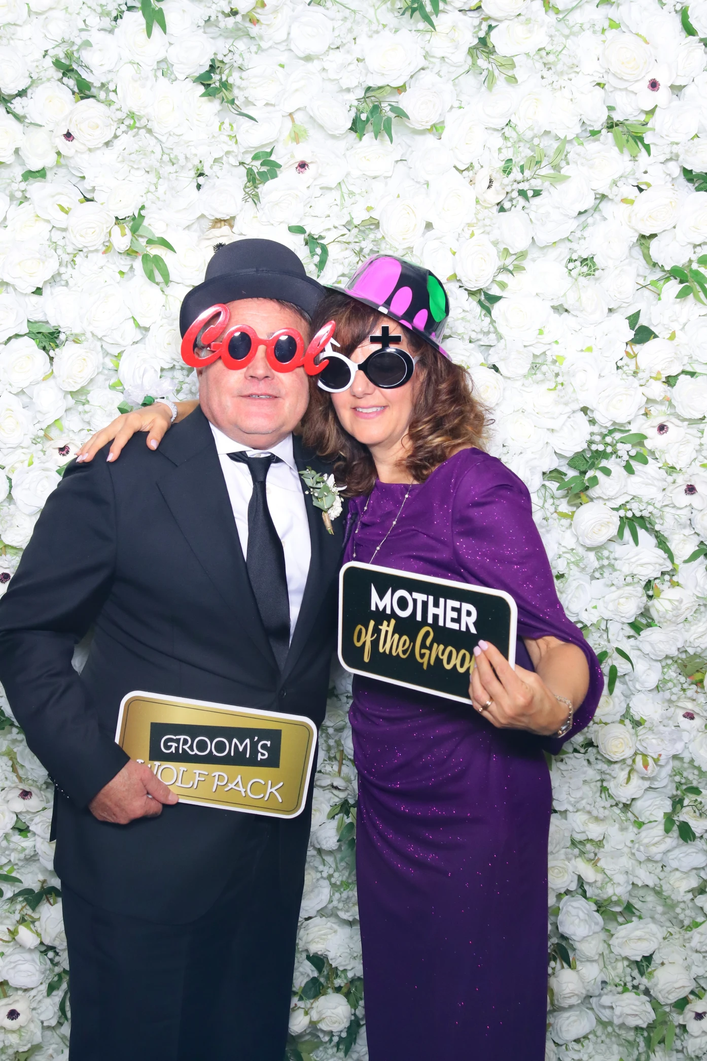 Man in black suit and woman in purple dress pose with funny hats, sunglasses, and signs reading 'Groom's Wolf Pack' and 'Mother of the Groom' in front of a white floral backdrop.