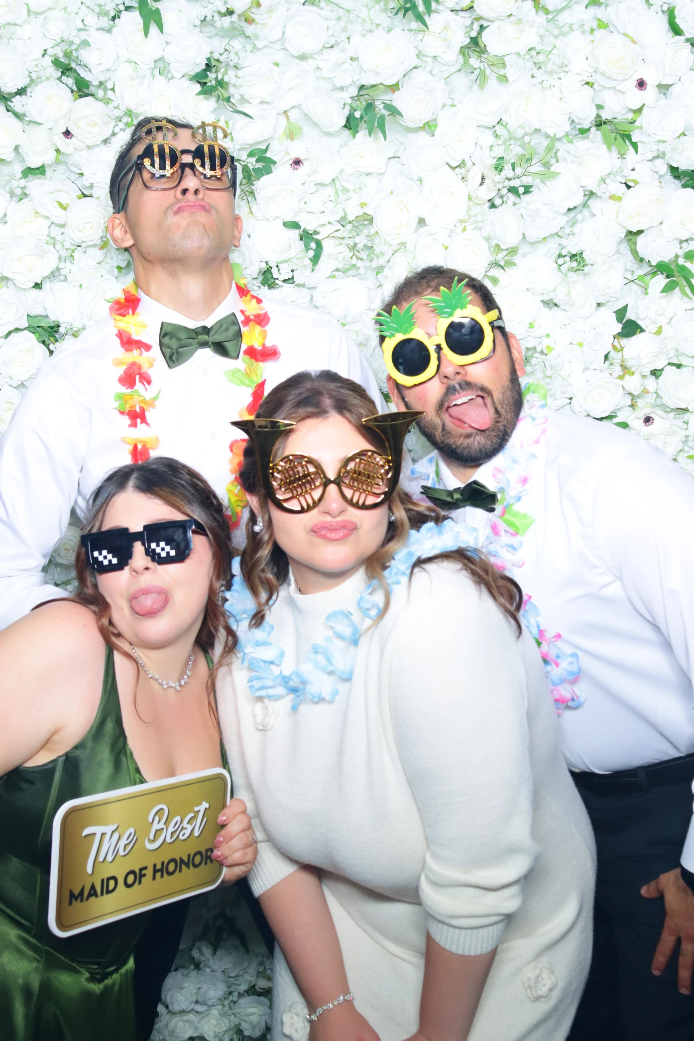 Four people in festive glasses and leis posing playfully in front of a white flower wall, one holding a sign that says 'The Best Maid of Honor.'
