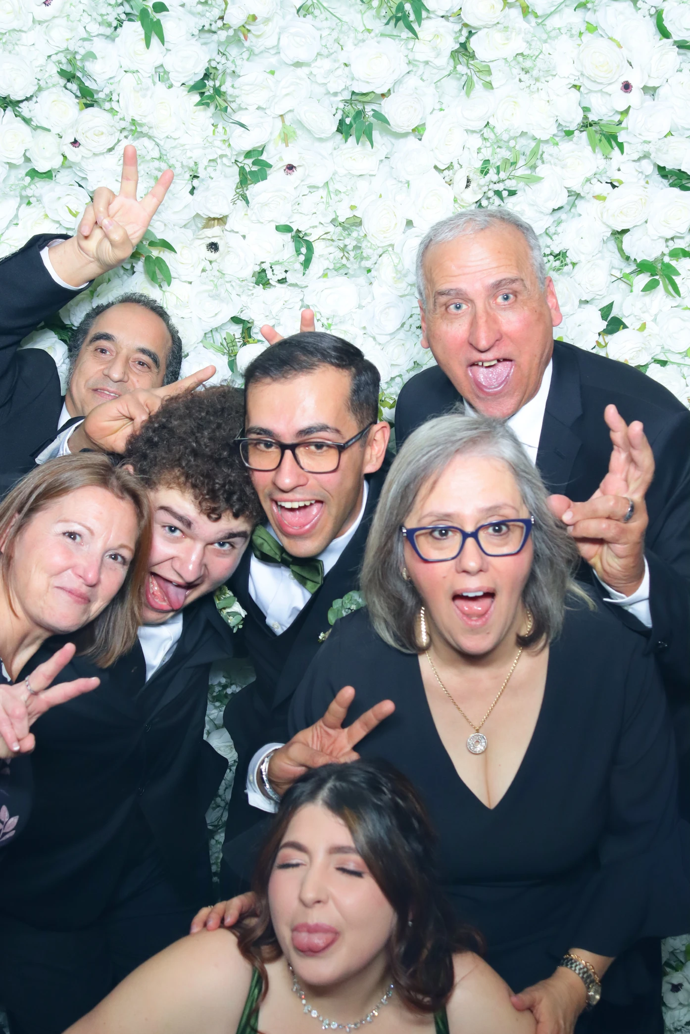 Group of seven people posing playfully in front of a white floral backdrop, making funny faces and peace signs.