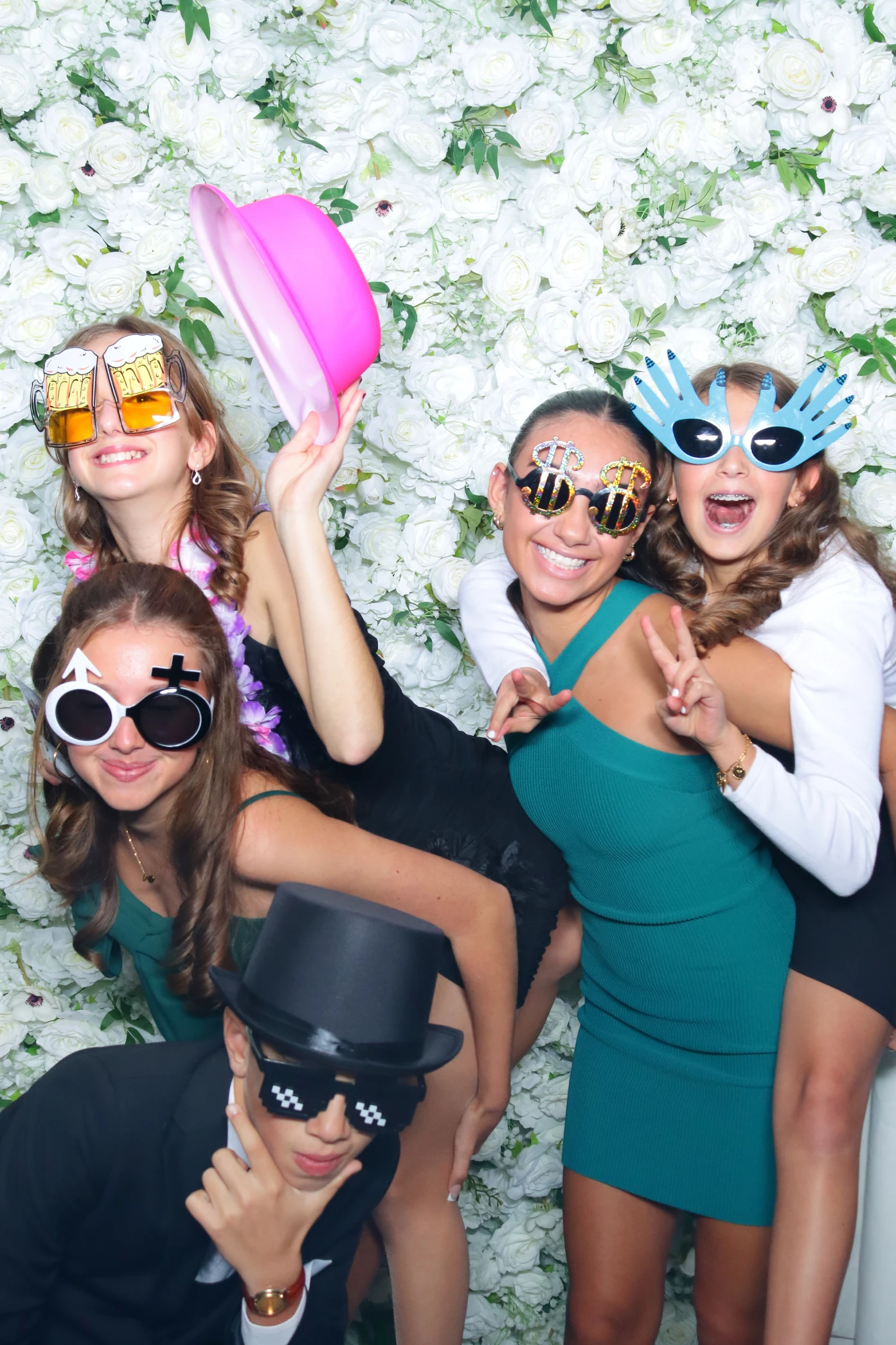 Group of five young people posing joyfully in front of a white flower wall, wearing playful novelty sunglasses and hats.
