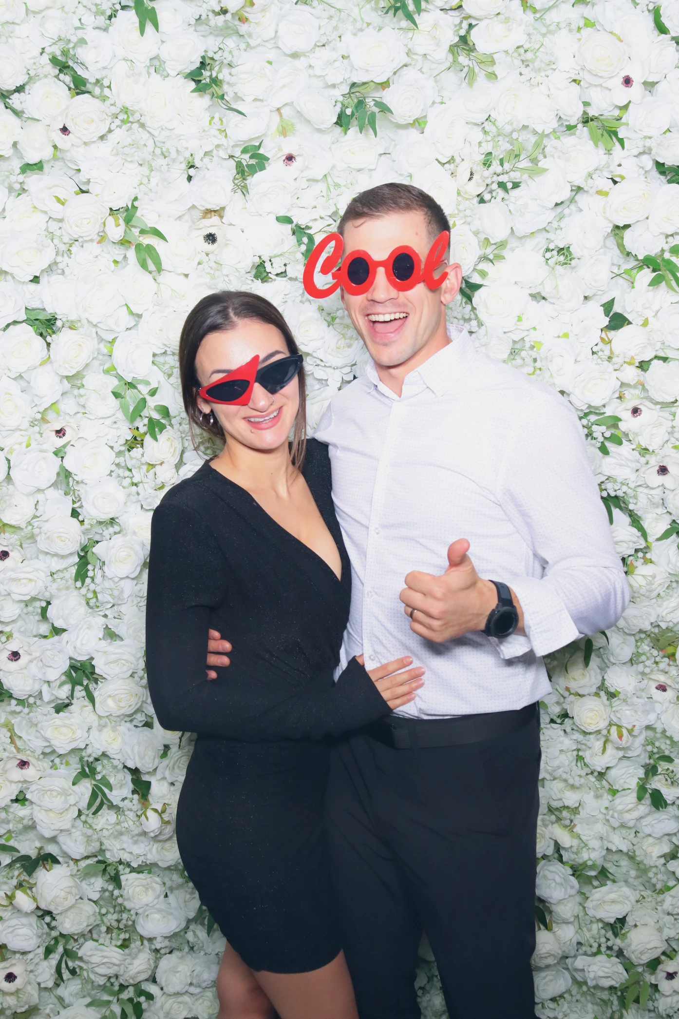 A smiling couple wearing playful red sunglasses, standing in front of a white rose flower wall; the man gives a thumbs up.