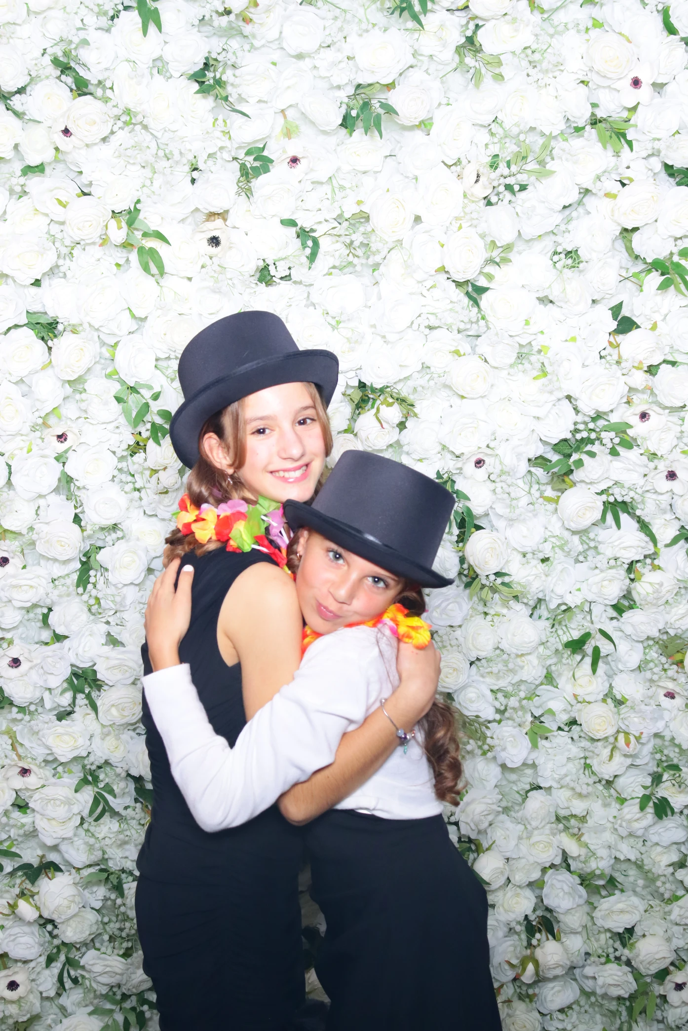 Two girls wearing black top hats and floral leis hugging in front of a white flower wall.