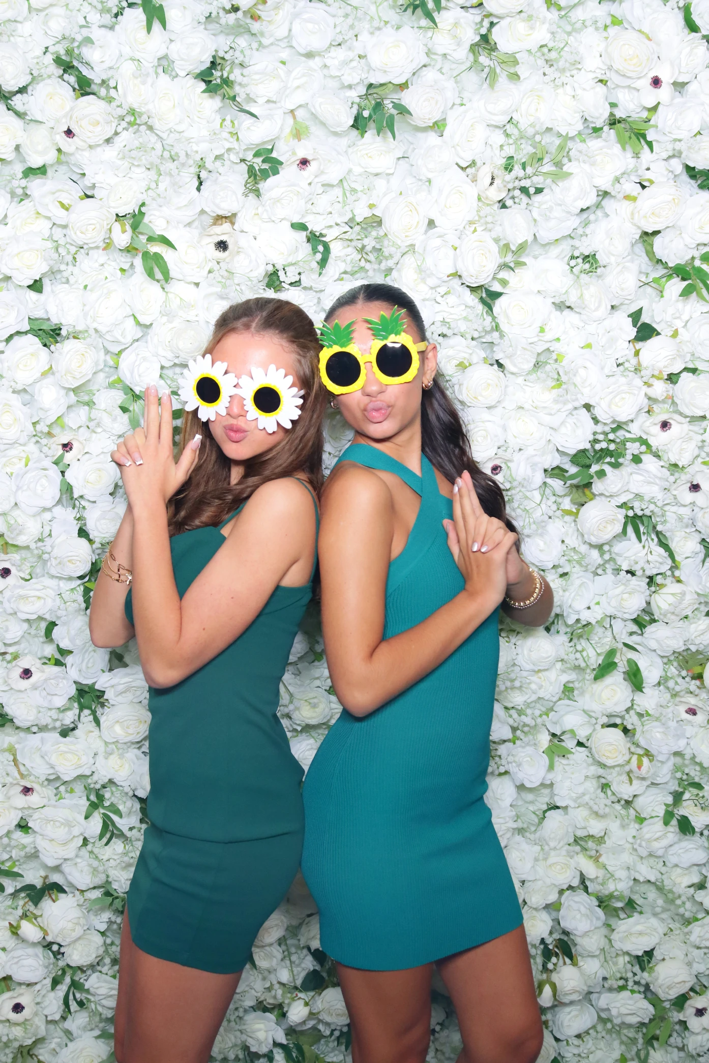 Two women in teal dresses wearing playful flower and pineapple sunglasses pose back-to-back against a white flower wall.