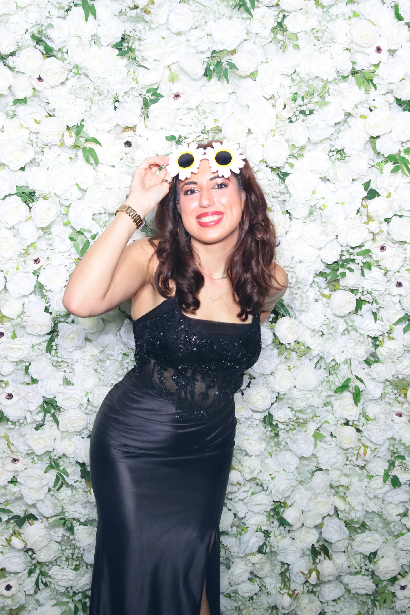 Smiling woman in a black dress wearing flower-shaped sunglasses posing in front of a white floral wall.