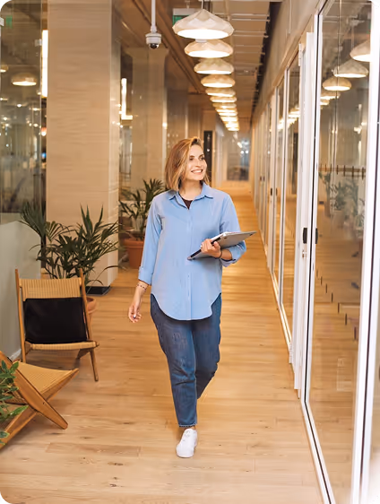 Woman in casual clothes holding a tablet walking down a brightly lit office corridor with wooden floors.