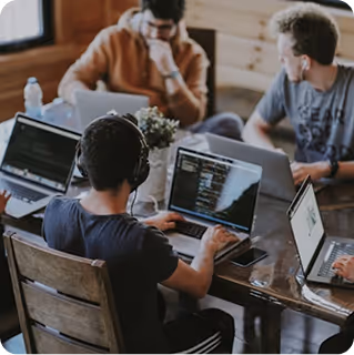 Three people working on laptops around a wooden table in a casual workspace.