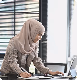 A woman wearing a beige hijab and plaid jacket writing in a notebook while using a laptop at a desk by a window with blinds.