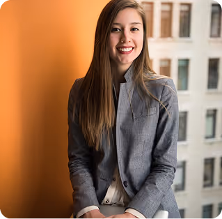 Smiling young woman with long brown hair wearing a gray blazer, seated indoors near a window with a blurred building in the background.