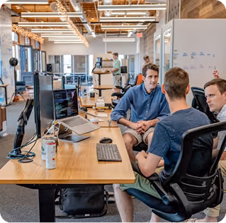 Three men engaged in a discussion around a desk with computer monitors and laptops in a modern open office space.