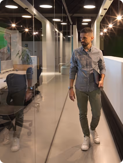 Man in casual attire walking in a modern office hallway holding a laptop.