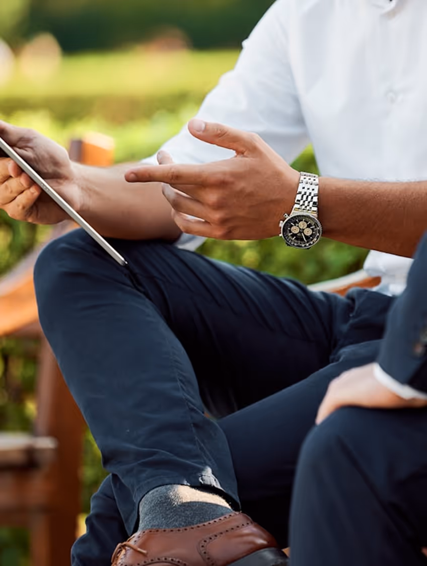 Man wearing a watch and brown dress shoes using a tablet while sitting outdoors, engaged in conversation.