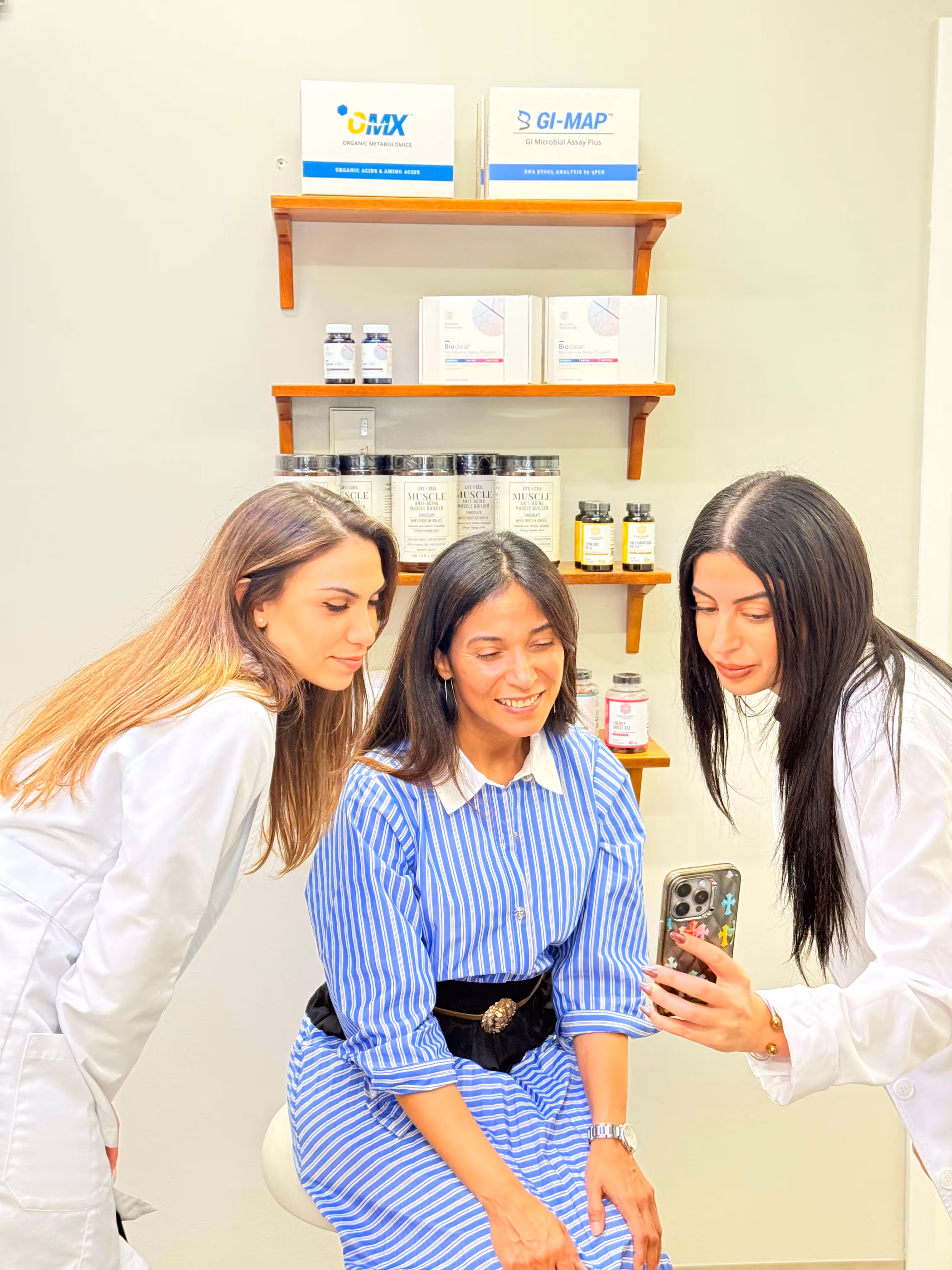 Three women, two in white coats, showing a phone screen to a smiling woman in a blue striped dress seated on a stool, with shelves of supplements in the background.