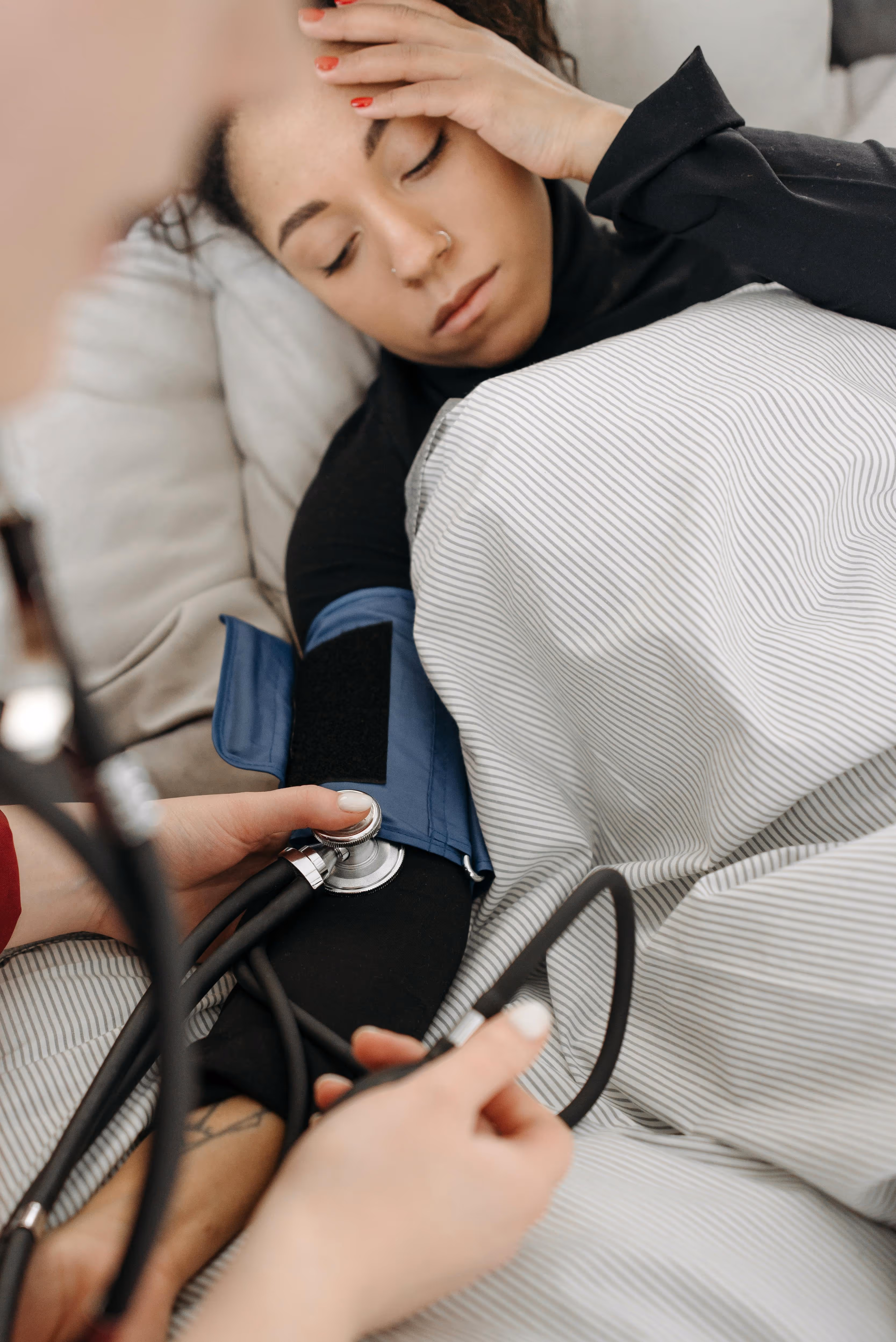 A person lying in bed with their eyes closed, having their blood pressure checked with a cuff and stethoscope.