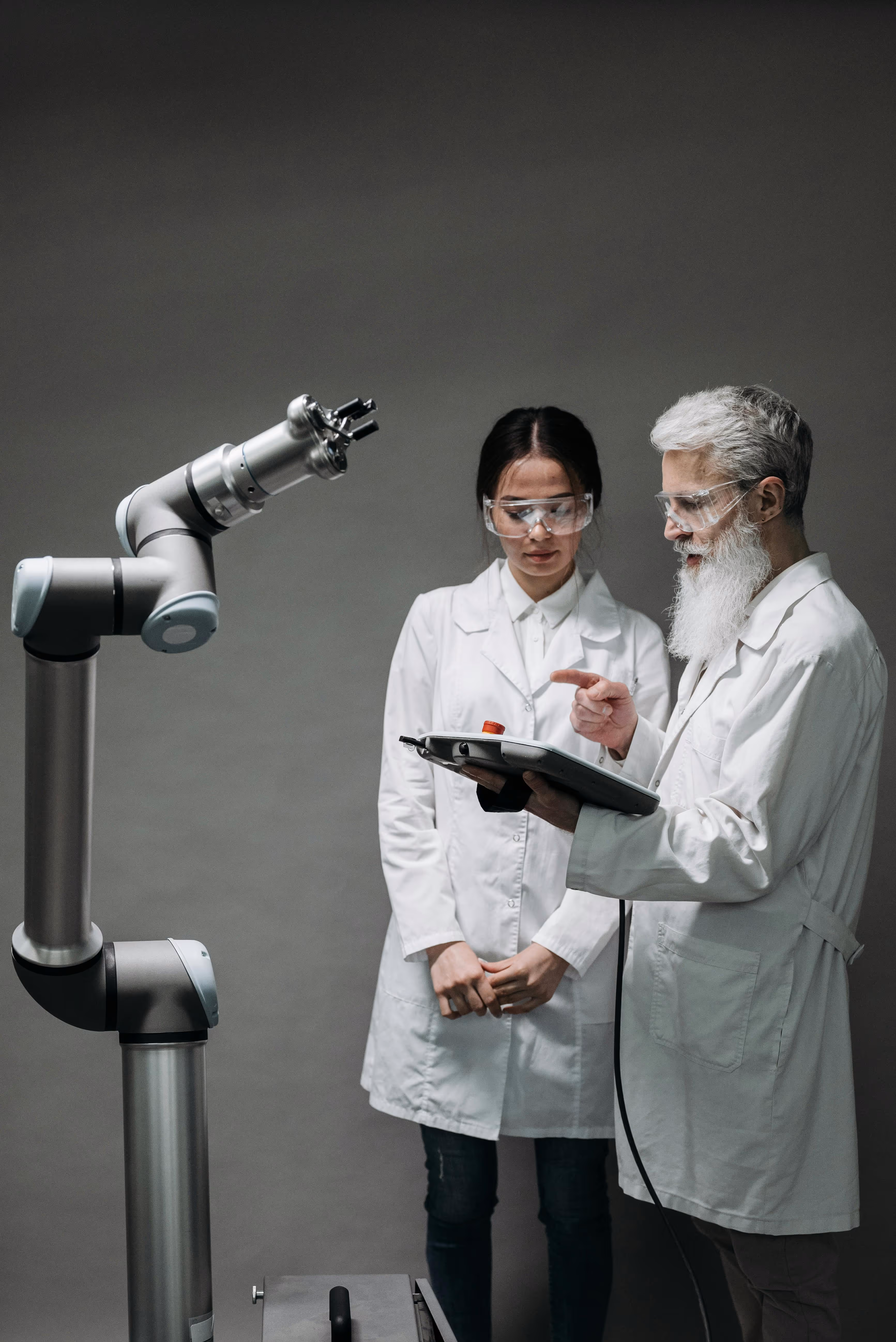 Two scientists in white lab coats and safety glasses operating a robotic arm using a control panel.