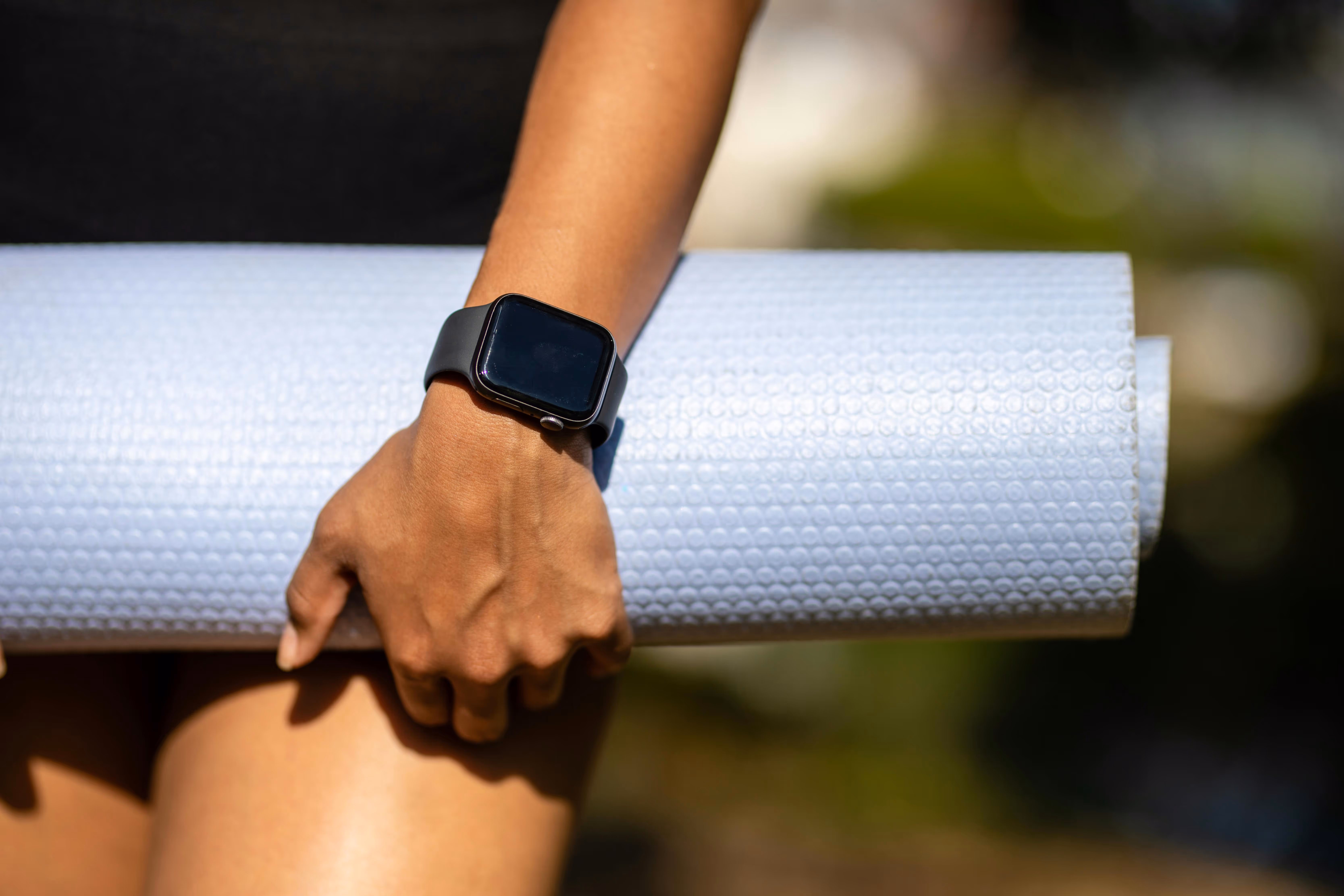 Person holding a rolled white yoga mat wearing a black smartwatch on their wrist.