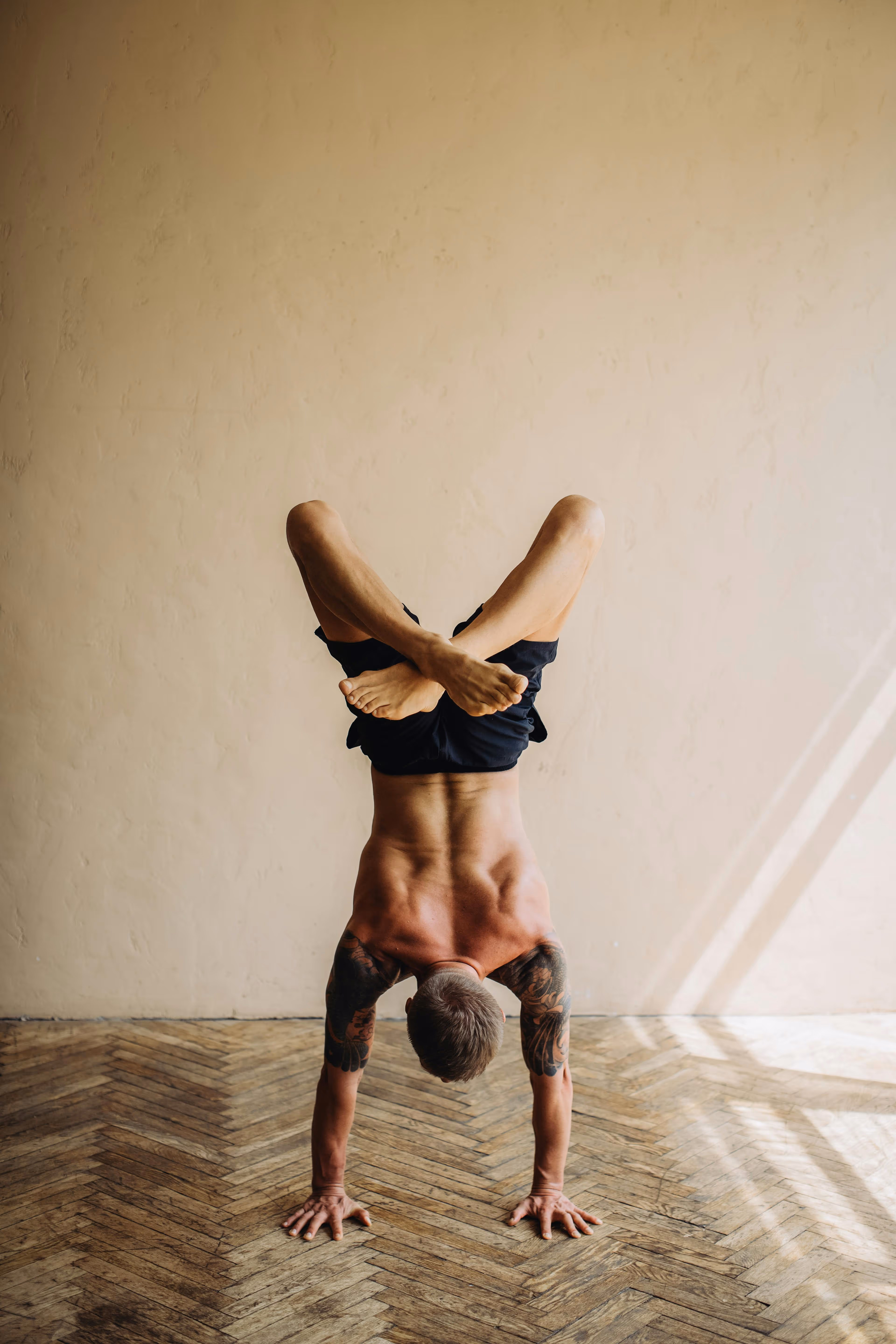 Tattooed man doing a yoga handstand with crossed legs on a wooden floor against a beige wall.