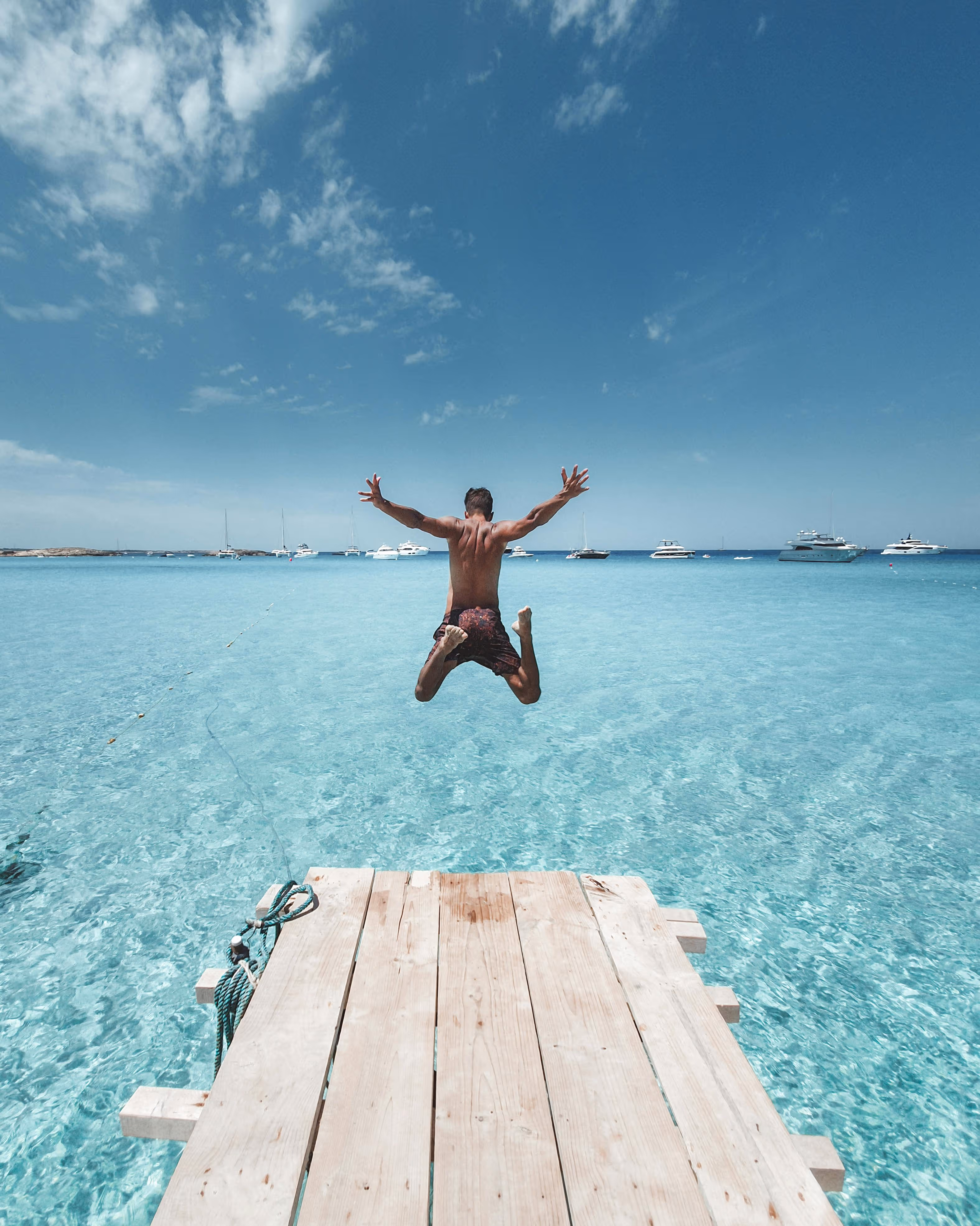Man jumping off a wooden dock into clear turquoise sea with boats and yachts in the distance under a partly cloudy blue sky.