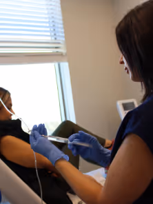 Healthcare worker wearing blue gloves prepares a syringe while a patient sits in a chair near a window.