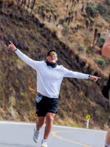 Runner in white shirt and black shorts crossing a road, celebrating with arms spread wide on a mountain path.
