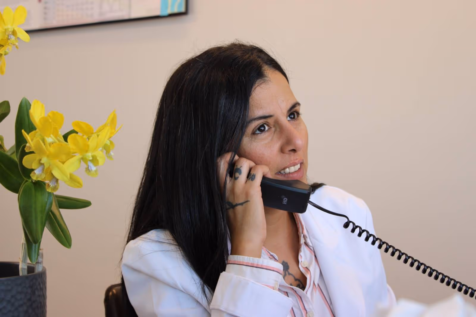 Woman with dark hair wearing a white coat talking on a corded phone, with yellow flowers in a pot nearby.