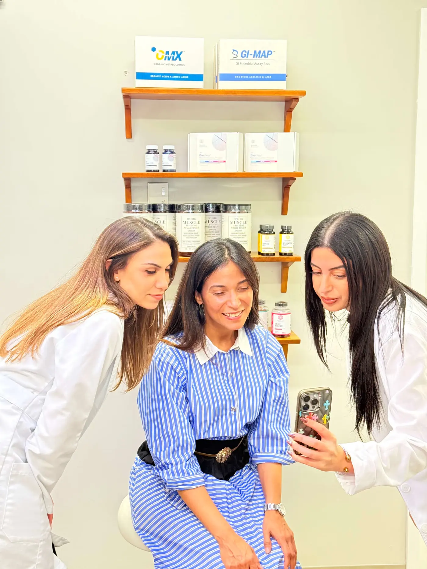 Two women in white lab coats showing a smartphone to a seated woman in a blue striped dress in a consultation room with shelves of supplements.