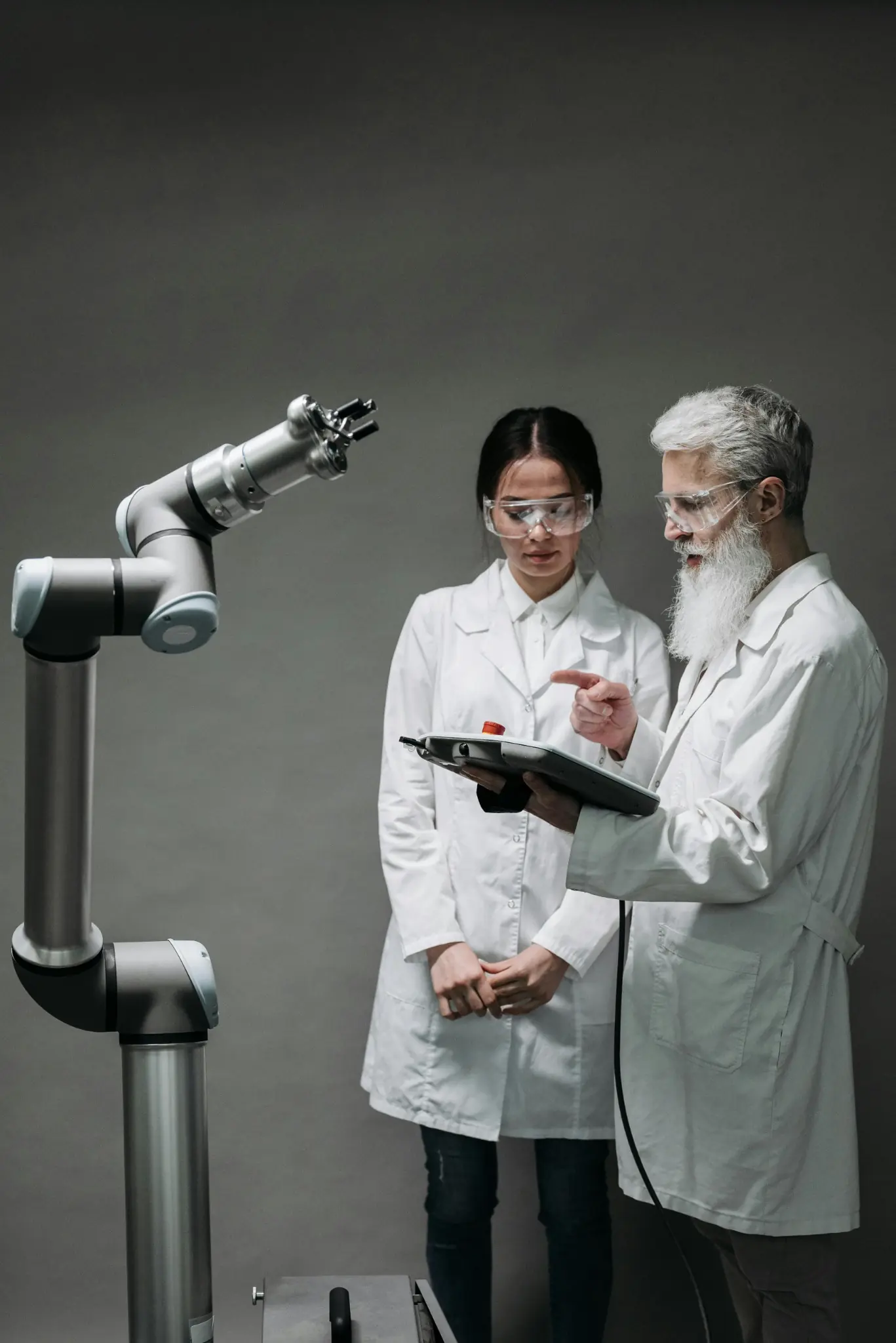 Two scientists in white lab coats and safety goggles examining a robotic arm using a control tablet.