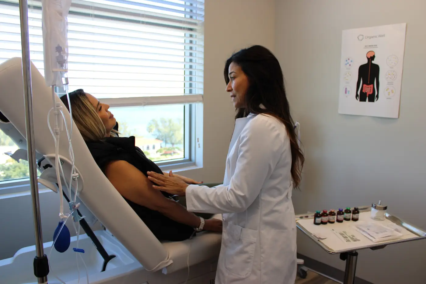 A female doctor in a white coat reassures a patient seated in a clinic chair with an IV drip next to her.