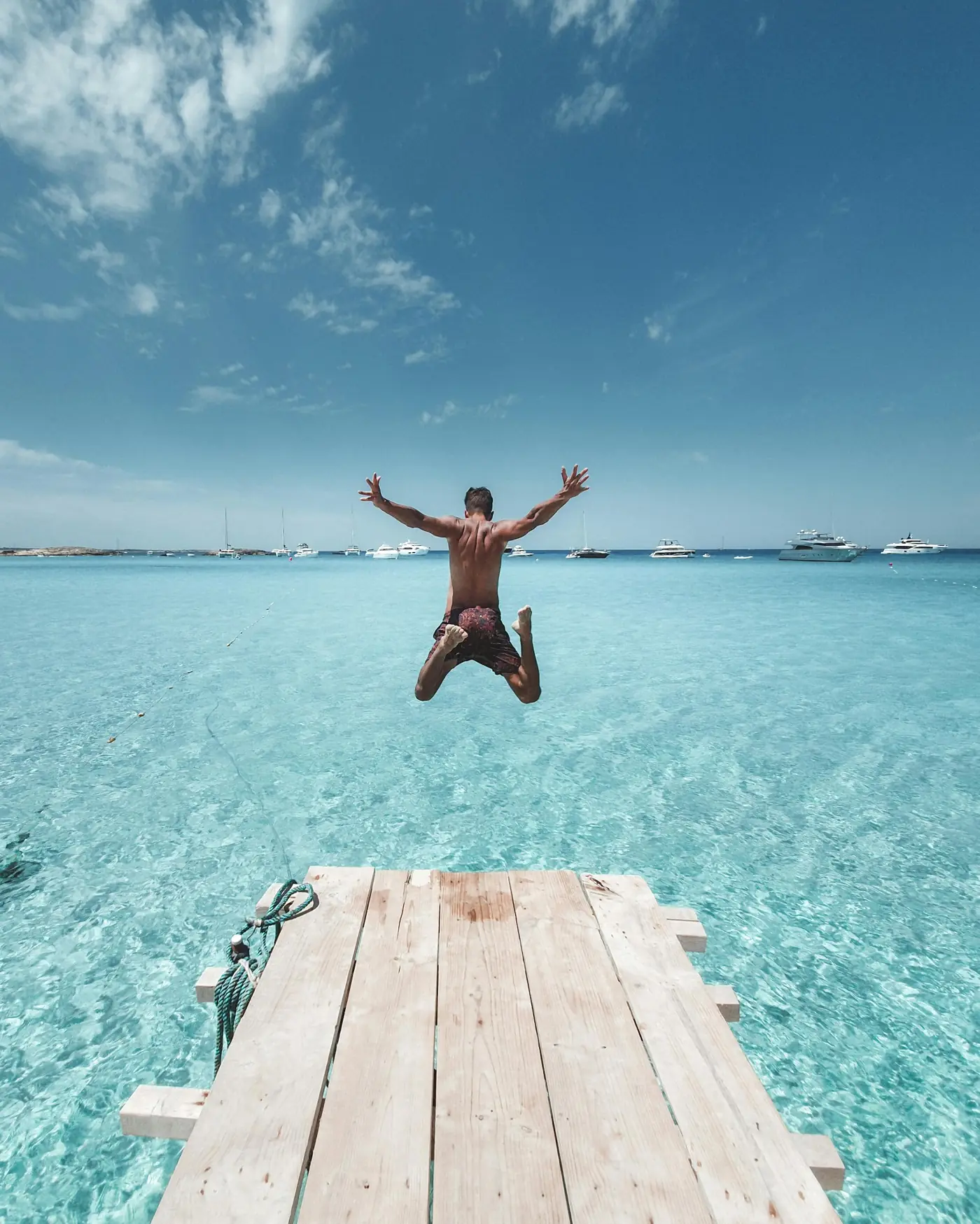 Man jumping off a wooden pier into clear, turquoise sea under a blue sky with scattered clouds and boats in the distance.