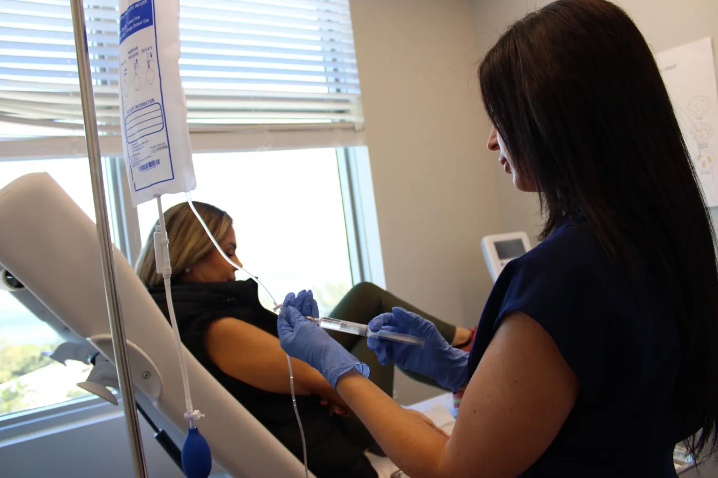 Healthcare professional wearing blue gloves prepares an IV drip for a patient reclining in a medical chair.