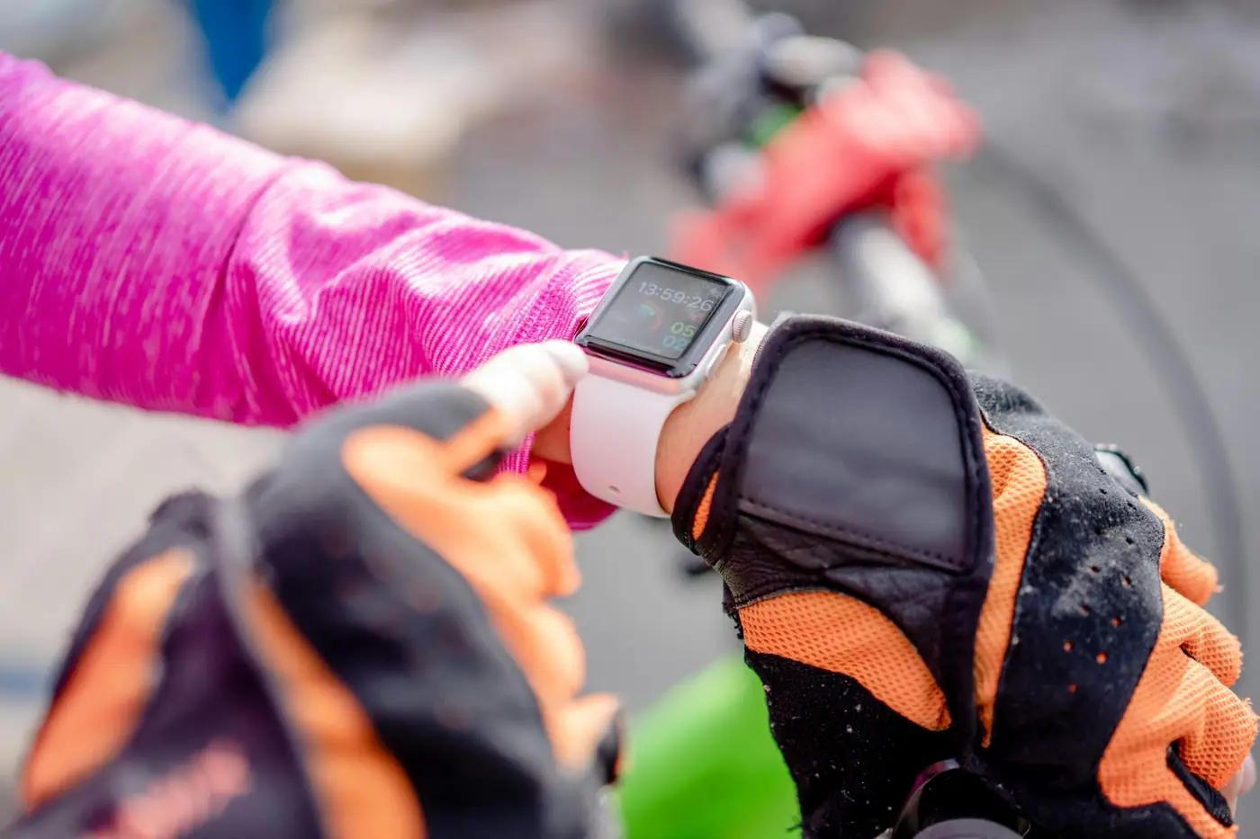 Person wearing orange and black cycling gloves and a pink jacket checking a white smartwatch while holding bicycle handlebars.