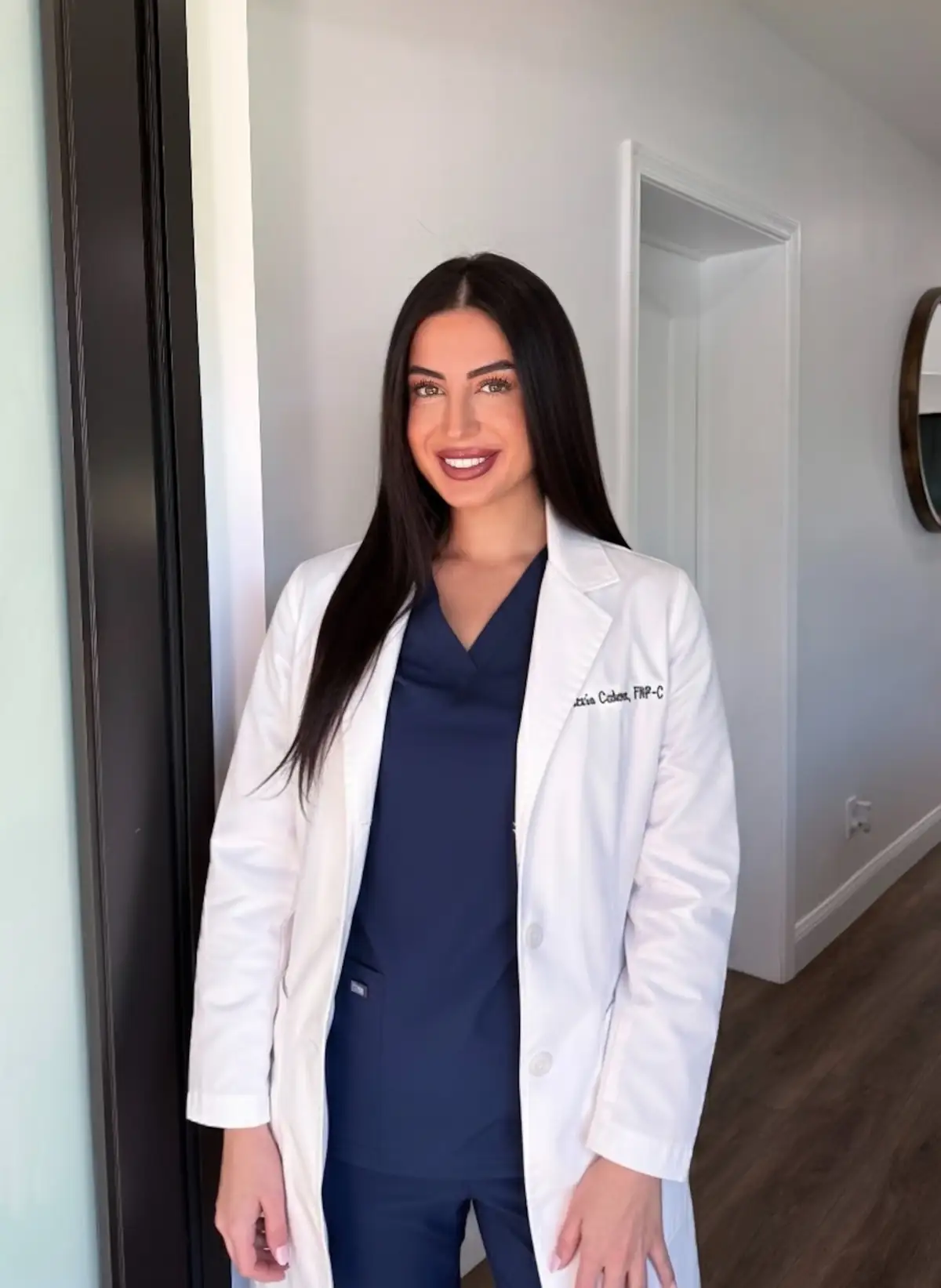 Smiling female doctor with long dark hair wearing a white lab coat over navy blue scrubs, standing indoors.