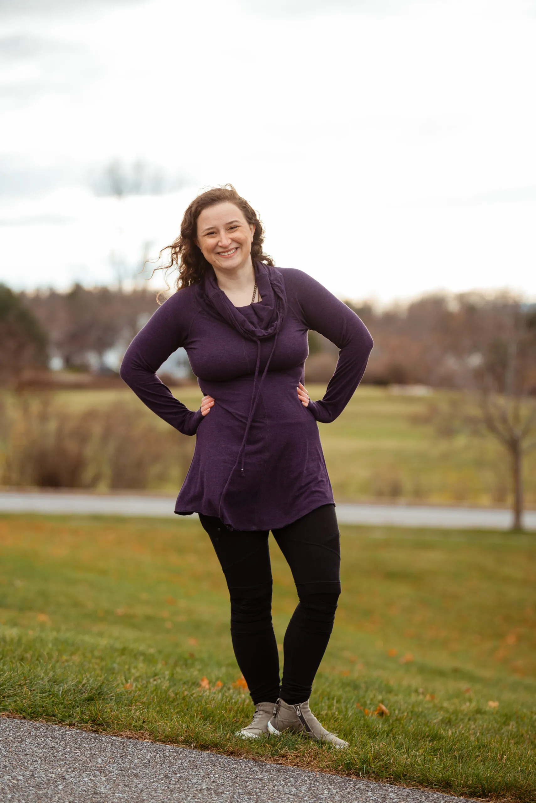 Erika Austin standing outdoors on grass next to a paved path with her hands on her hips, wearing a purple long-sleeve top, black leggings, and beige ankle boots.