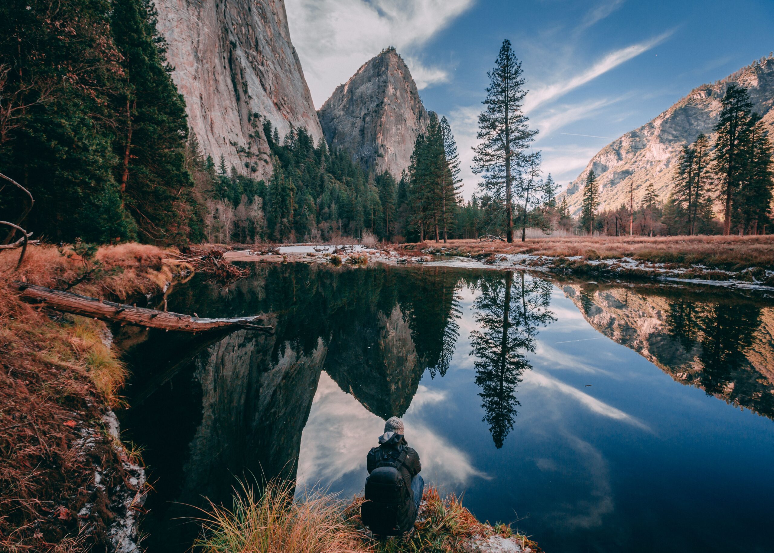 Person sitting near a calm river reflecting pine trees and mountains under a partly cloudy sky.