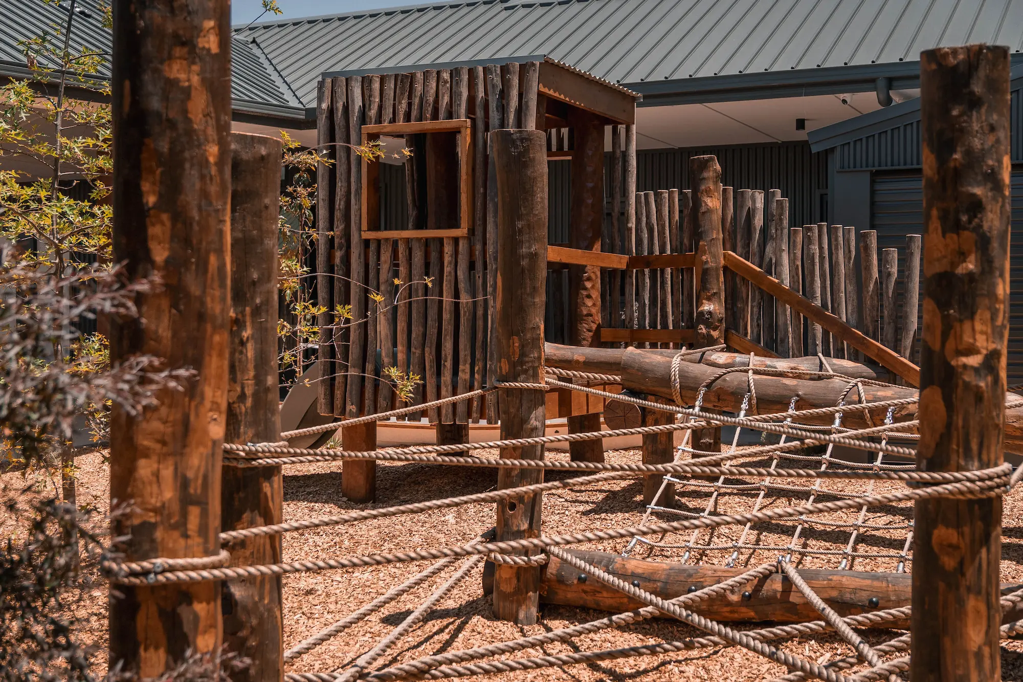 Wooden playground structure featuring a climbing frame, ropes, and natural surroundings with soft ground cover. Ideal for children's play.