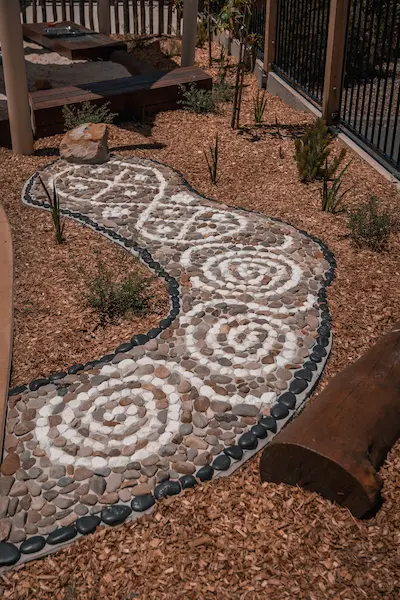 Stone pathway with spiral patterns, surrounded by gravel and plants, in a landscaped outdoor area.