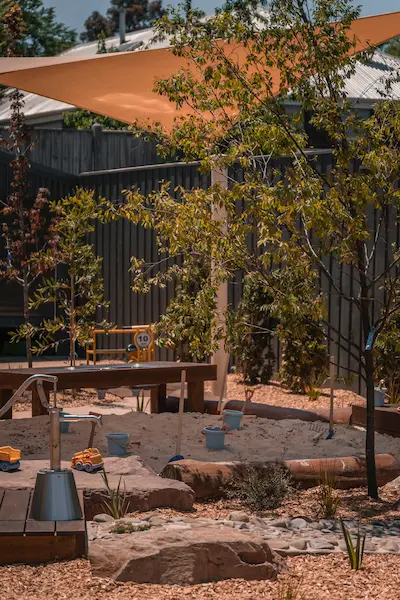 A playground with a sand area, surrounded by trees and rocks, features a shade sail overhead and children's toys scattered around.