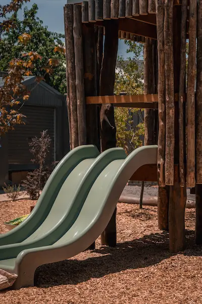 Three green playground slides attached to a wooden play structure, surrounded by wood chips and greenery in a playground setting.