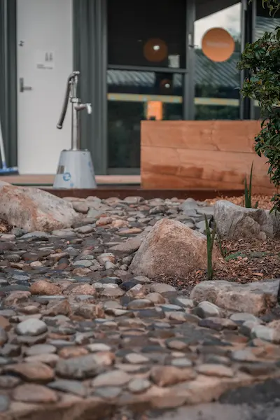 A rocky pathway leads to a modern entrance with a wooden bench and greenery beside the door.