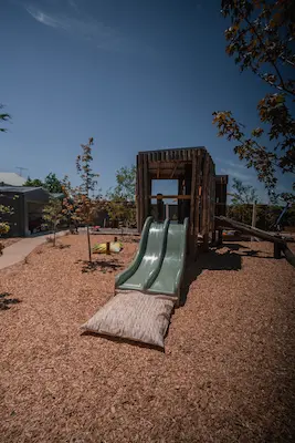 Playground with a wooden structure featuring two green slides on a bed of mulch, surrounded by young trees and clear blue sky.