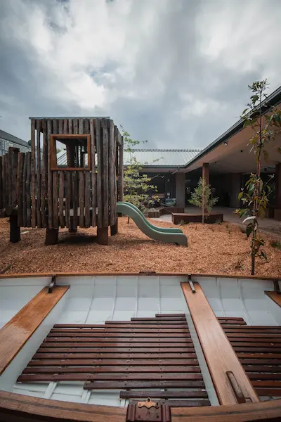 Playground with a wooden playhouse and a green slide, surrounded by mulch and young trees, under a cloudy sky.