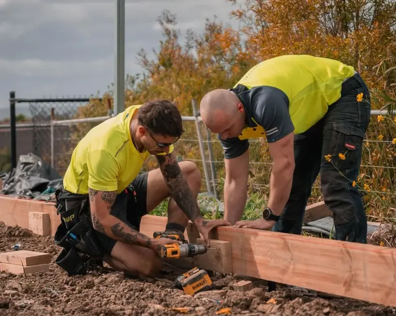 Two men in yellow work shirts are assembling wooden beams on a construction site, using a power drill and focused on their task.