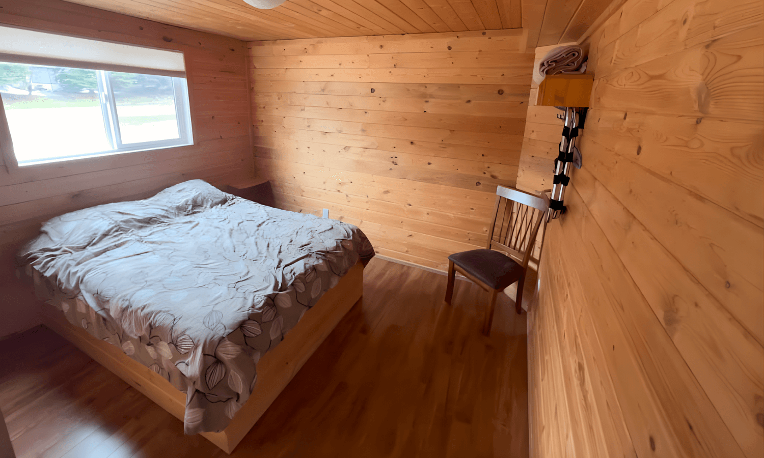 : Cabin bedroom with wood-paneled walls, floral comforter bed, and natural window light.