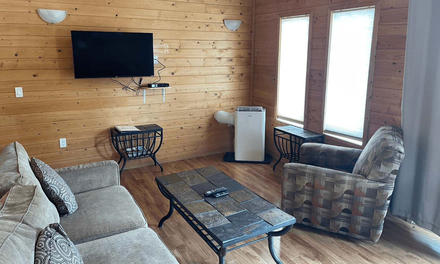 living room with wood-paneled walls, tan sofa, patterned armchair, stone-topped coffee table, and wall-mounted TV