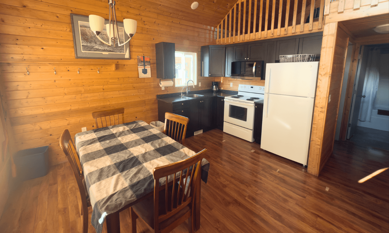 cabin interior with wooden dining table, checkered tablecloth, compact kitchen, and loft above.